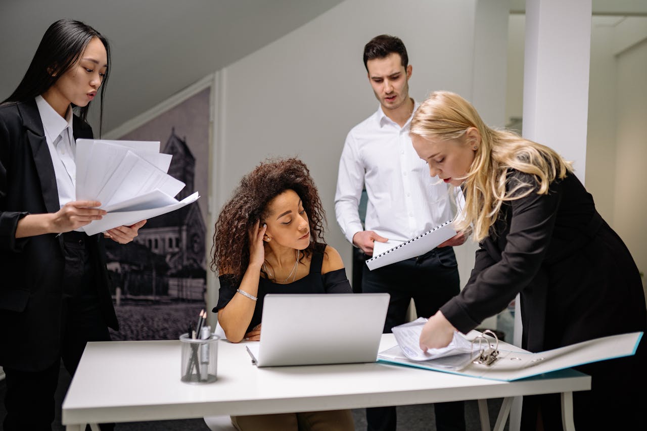 who-we-are A diverse group of coworkers brainstorming over paperwork and laptops in a corporate office setting.